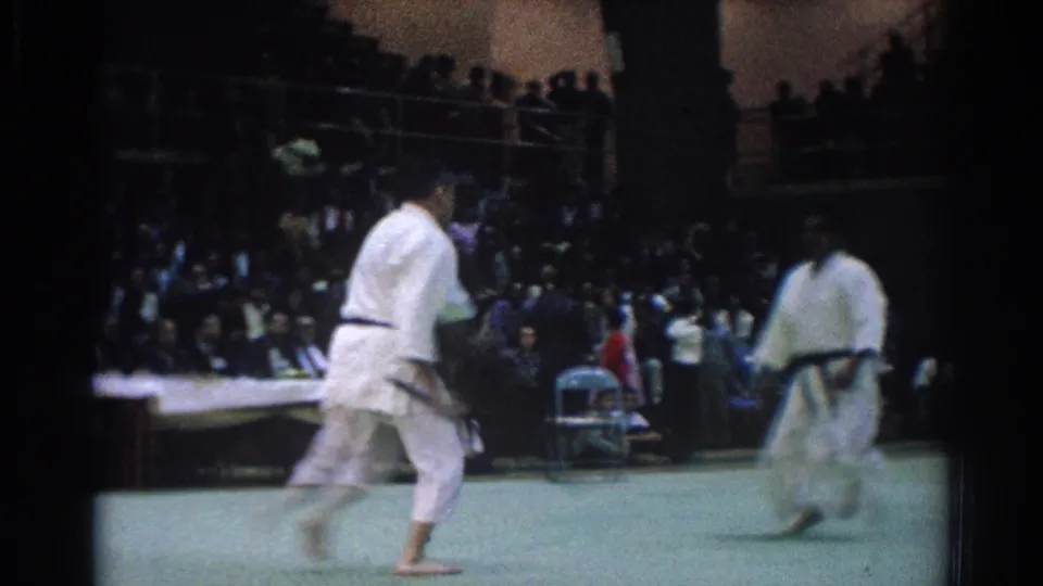 In 1966 West Covina footage, two black belt judo fighters in white gi square off on a green mat inside a crowded indoor arena.