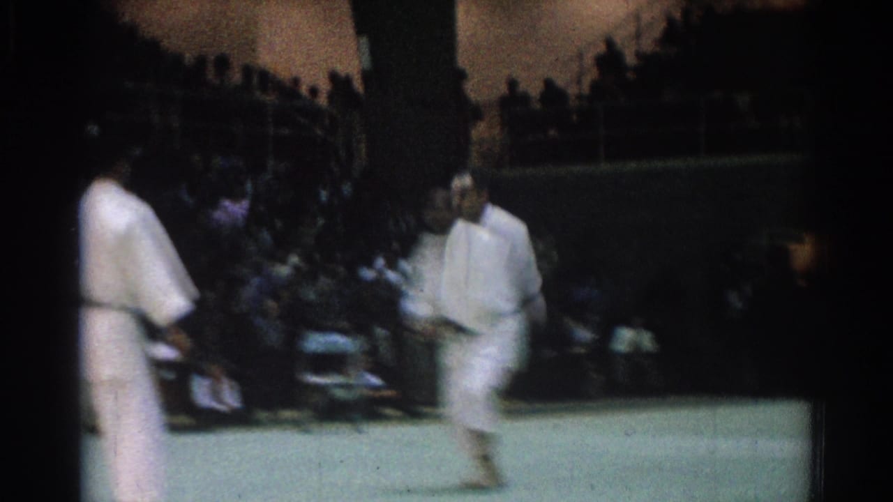 Two black belt judo competitors move across the green mat during a 1966 indoor match in West Covina, with spectators behind them.