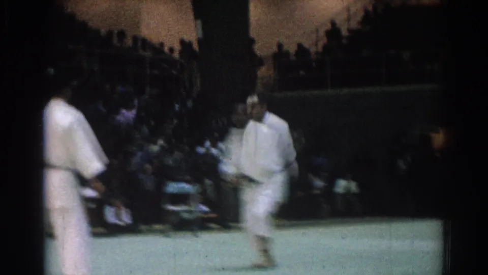 Two black belt judo competitors move across the green mat during a 1966 indoor match in West Covina, with spectators behind them.