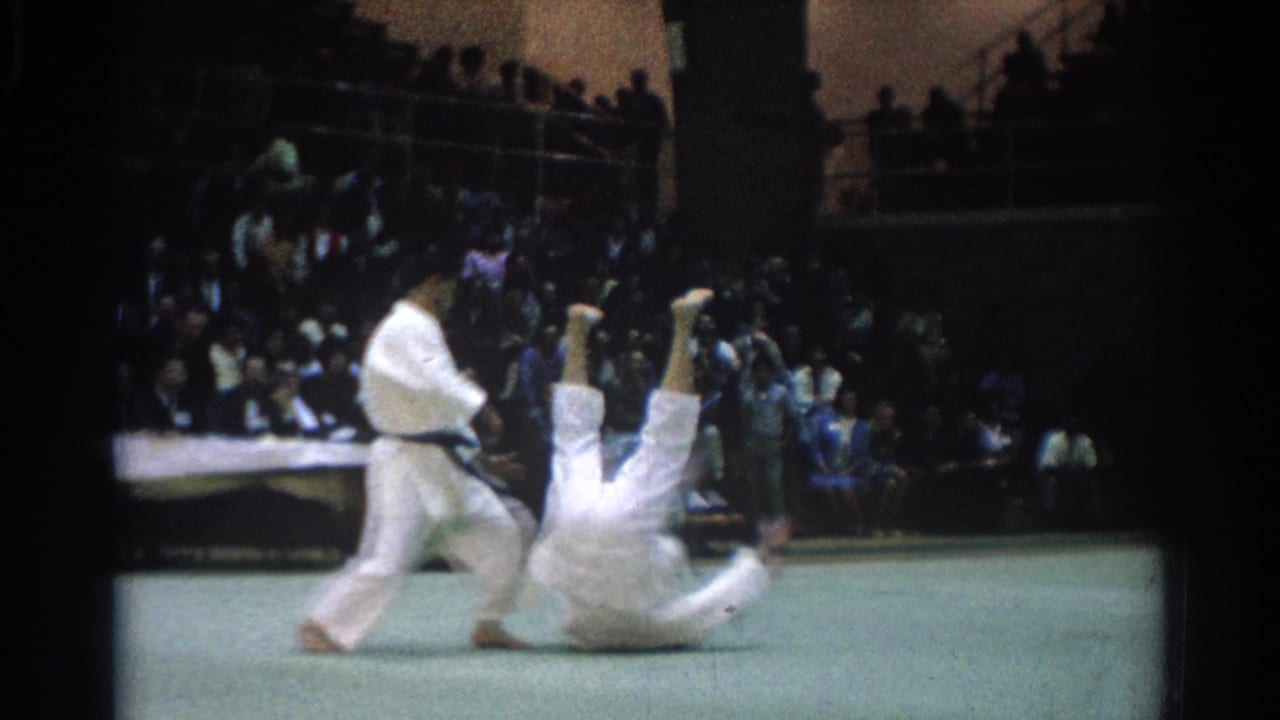 A judo throw is captured mid-action, with one fighter upright and the other landing backward on the green mat before the crowd.