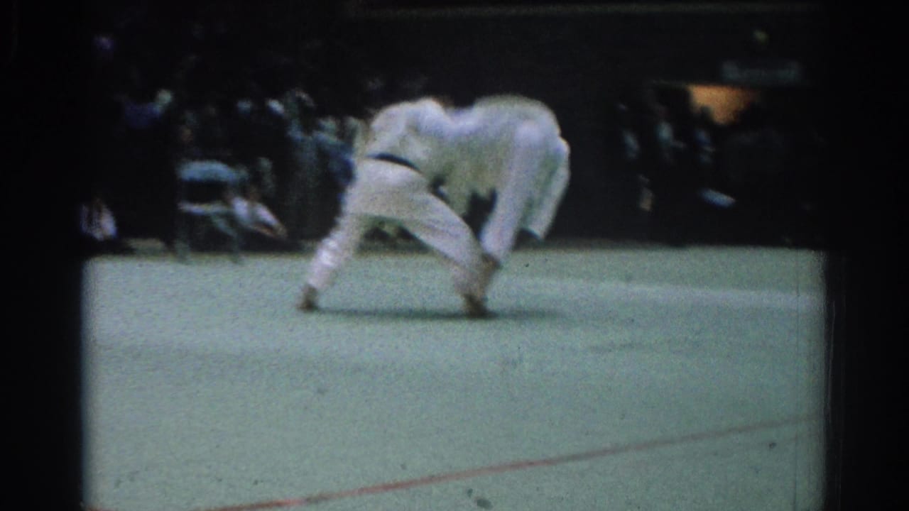 The match continues with both judo fighters bent low in a grappling exchange on the green mat inside the crowded arena.