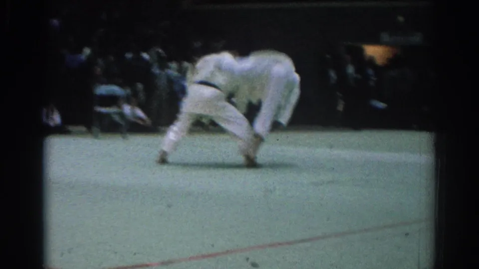 The match continues with both judo fighters bent low in a grappling exchange on the green mat inside the crowded arena.