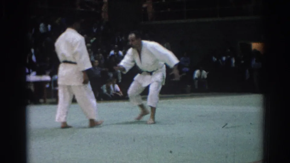One adult judo performer crouches low with an extended hand while the other stands ready on the mat in the 1966 home movie.