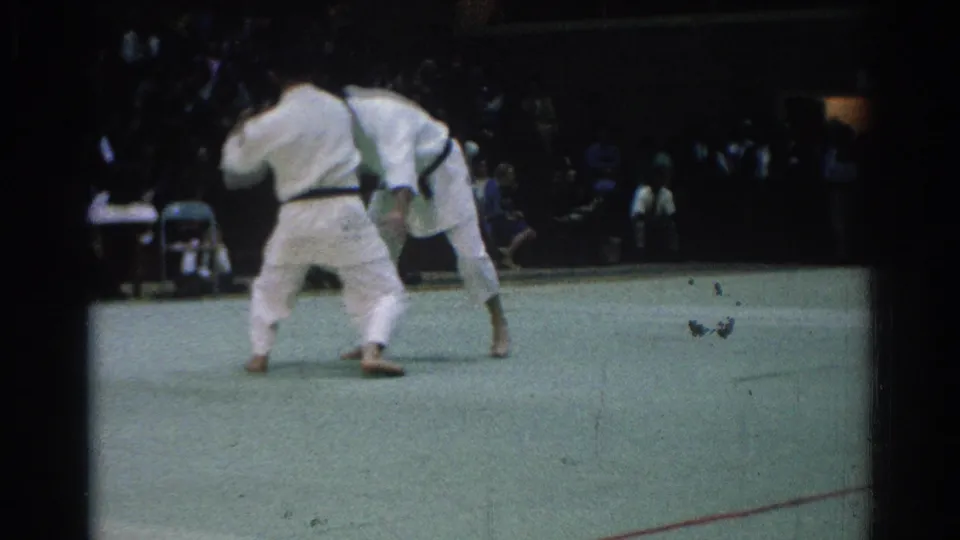 The two judo performers lean into a close grip, with spectators visible behind them in the West Covina community setting.