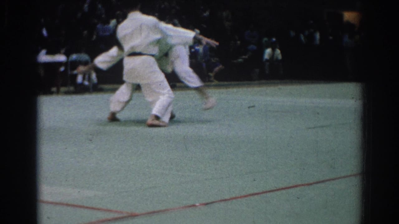 Two martial artists in white uniforms grapple on a green indoor mat.