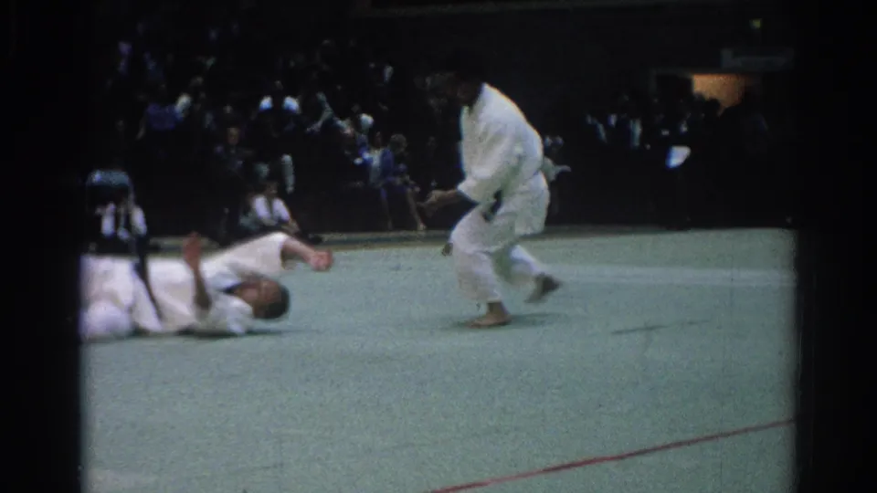 One adult competitor falls onto the green mat as another stands over him during the West Covina martial arts sequence.
