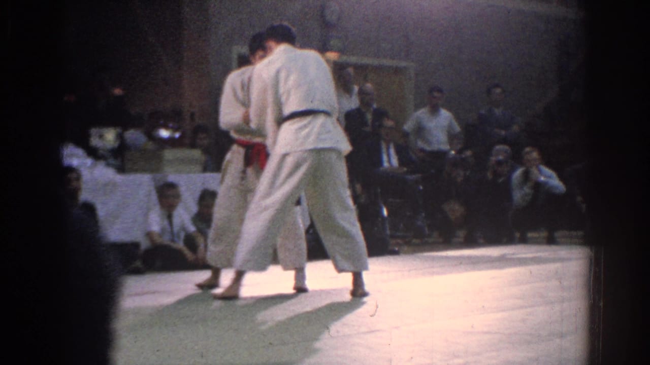 1966 West Covina tournament footage shows two martial artists in white gi, one with a red belt and one with a dark belt, grappling on an indoor mat.