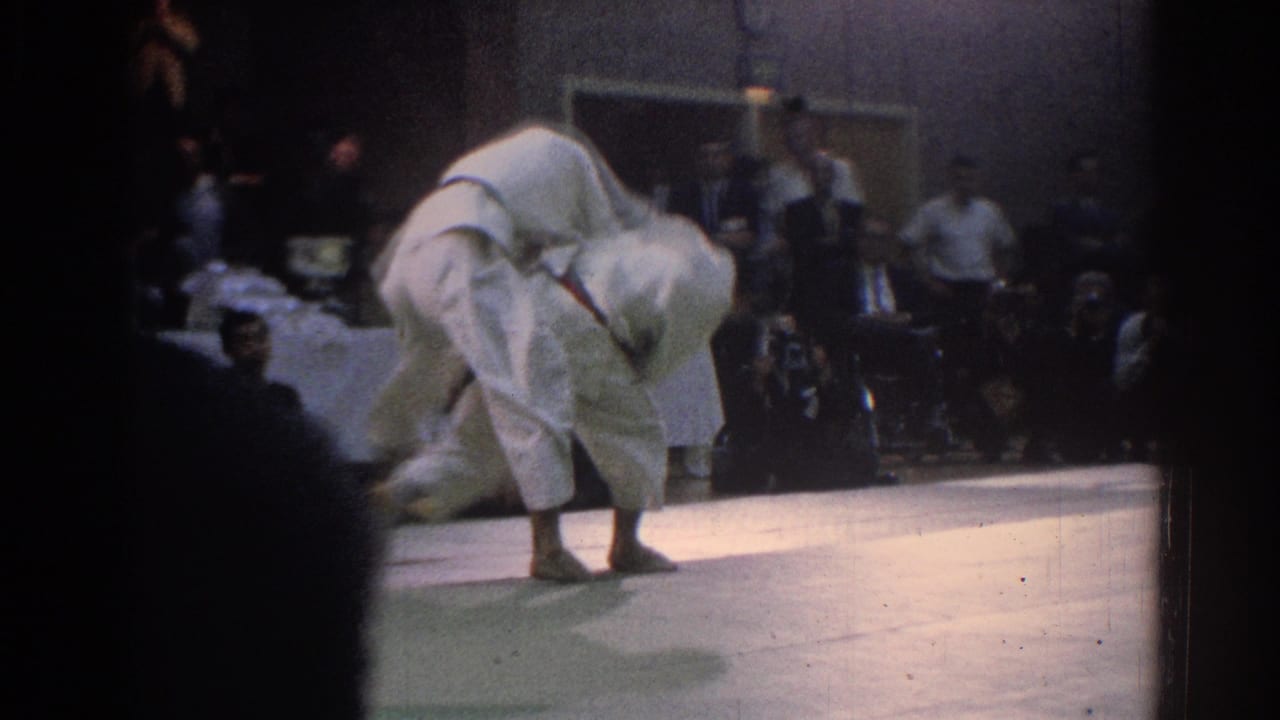 A competitor in a white gi bends into a throw or hold during the 1966 West Covina indoor martial arts tournament, with spectators seated behind the mat.