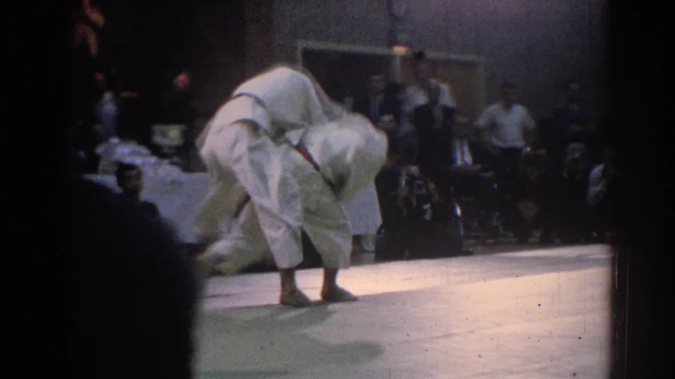 A competitor in a white gi bends into a throw or hold during the 1966 West Covina indoor martial arts tournament, with spectators seated behind the mat.