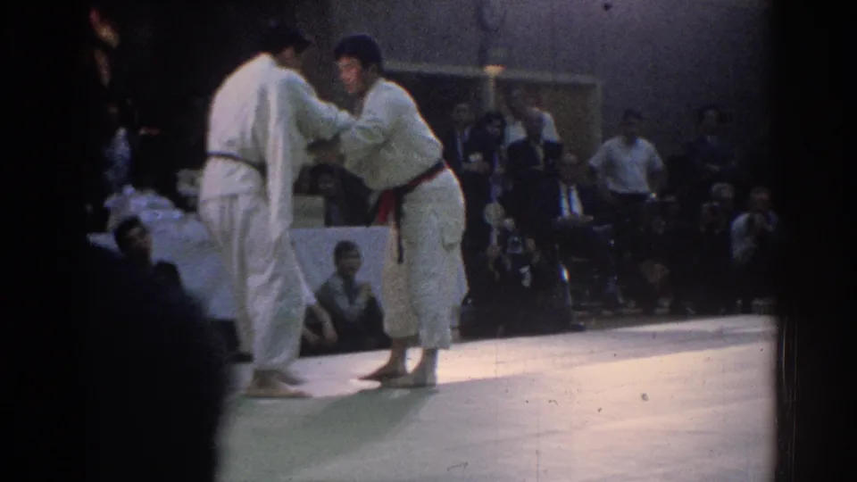 Two martial artists in white gi reset or grip near the center of the mat during a 1966 West Covina indoor tournament, with spectators visible in the background.