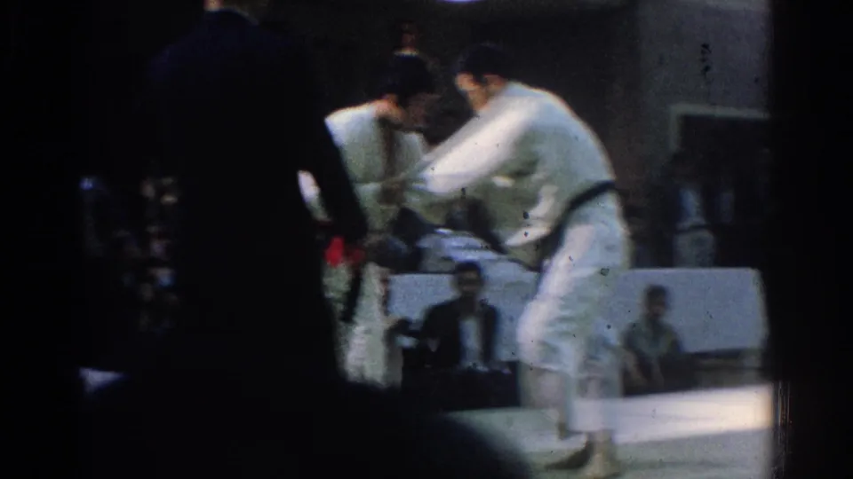 A white-gi competitor with a dark belt appears to pull or turn an opponent during the West Covina martial arts match, partly obscured by foreground silhouettes.