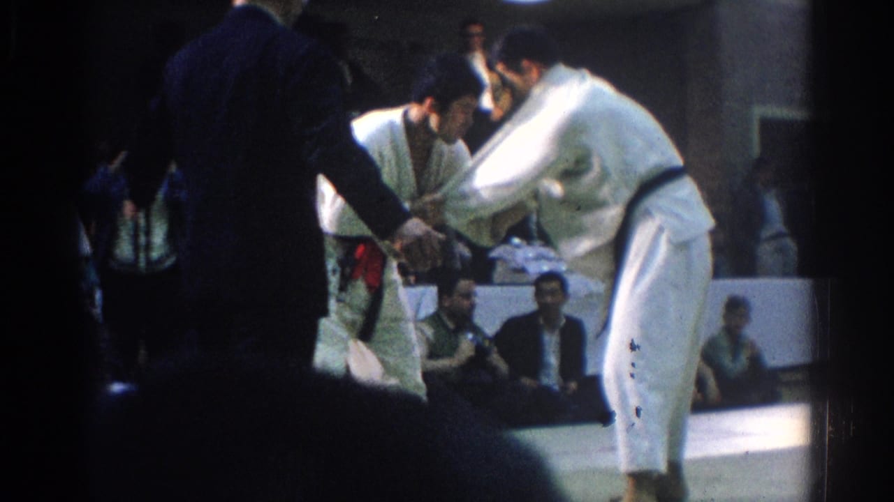 Two judoka in white gi grapple on an indoor mat while spectators sit behind them.