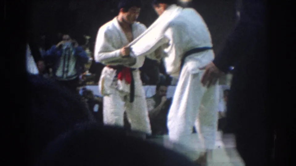 1966 West Covina frame with two judo competitors facing each other under indoor lighting, with audience members in the background.
