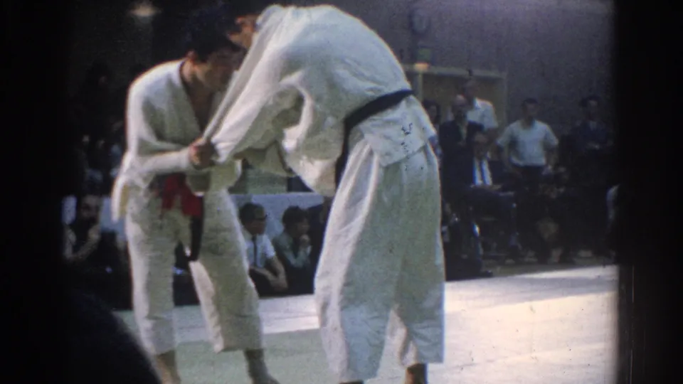 1966 West Covina frame showing the athletes locked in close grips during a judo exchange, filmed from a spectator position.