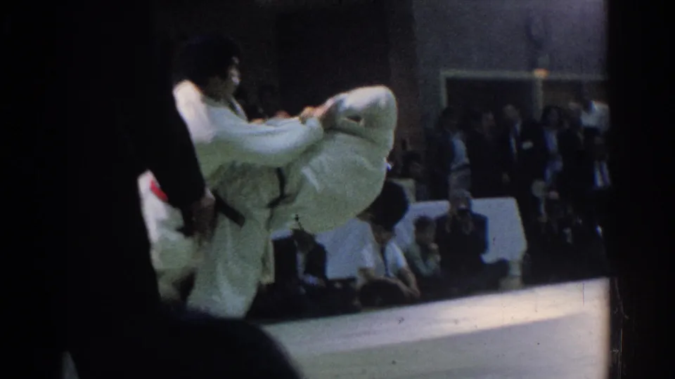 In 1966 West Covina footage, two judo competitors in white gis grapple on a mat with spectators seated behind them.