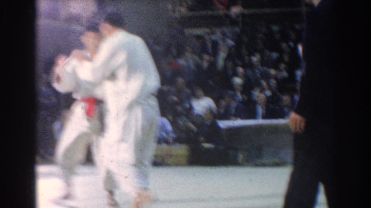 Two judo competitors stand close together on a mat while spectators watch.