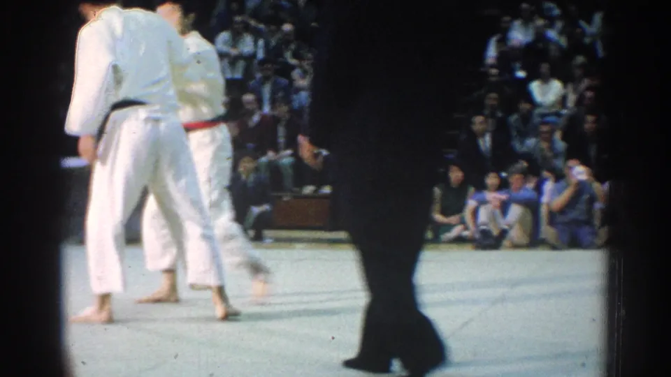 A handheld 8mm frame shows two judo competitors on a mat as a foreground figure partly obscures the 1966 West Covina scene.