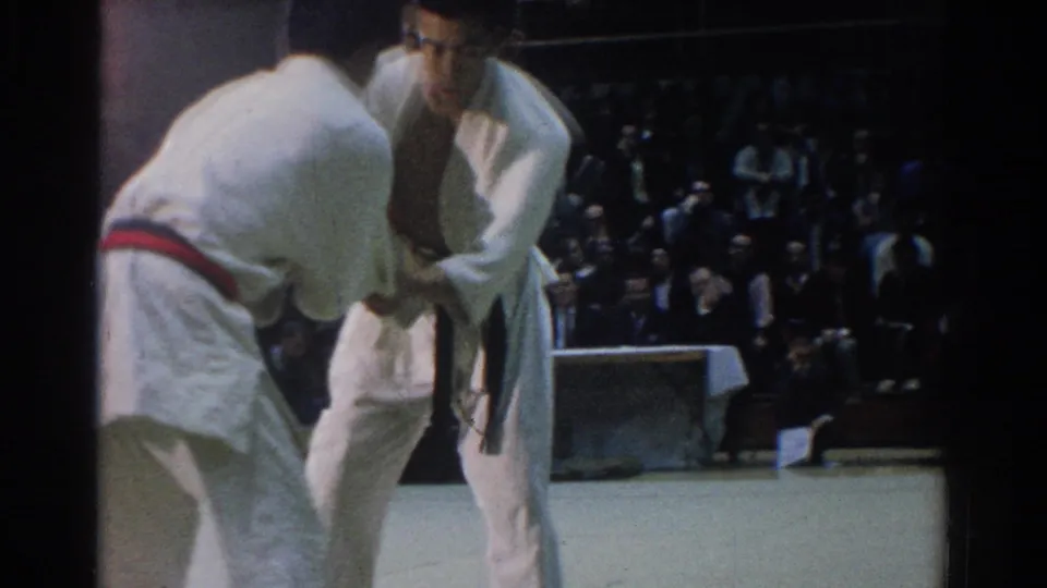 The competitors remain locked in close grip on the mat, with the seated audience visible in the dim 1966 West Covina hall.