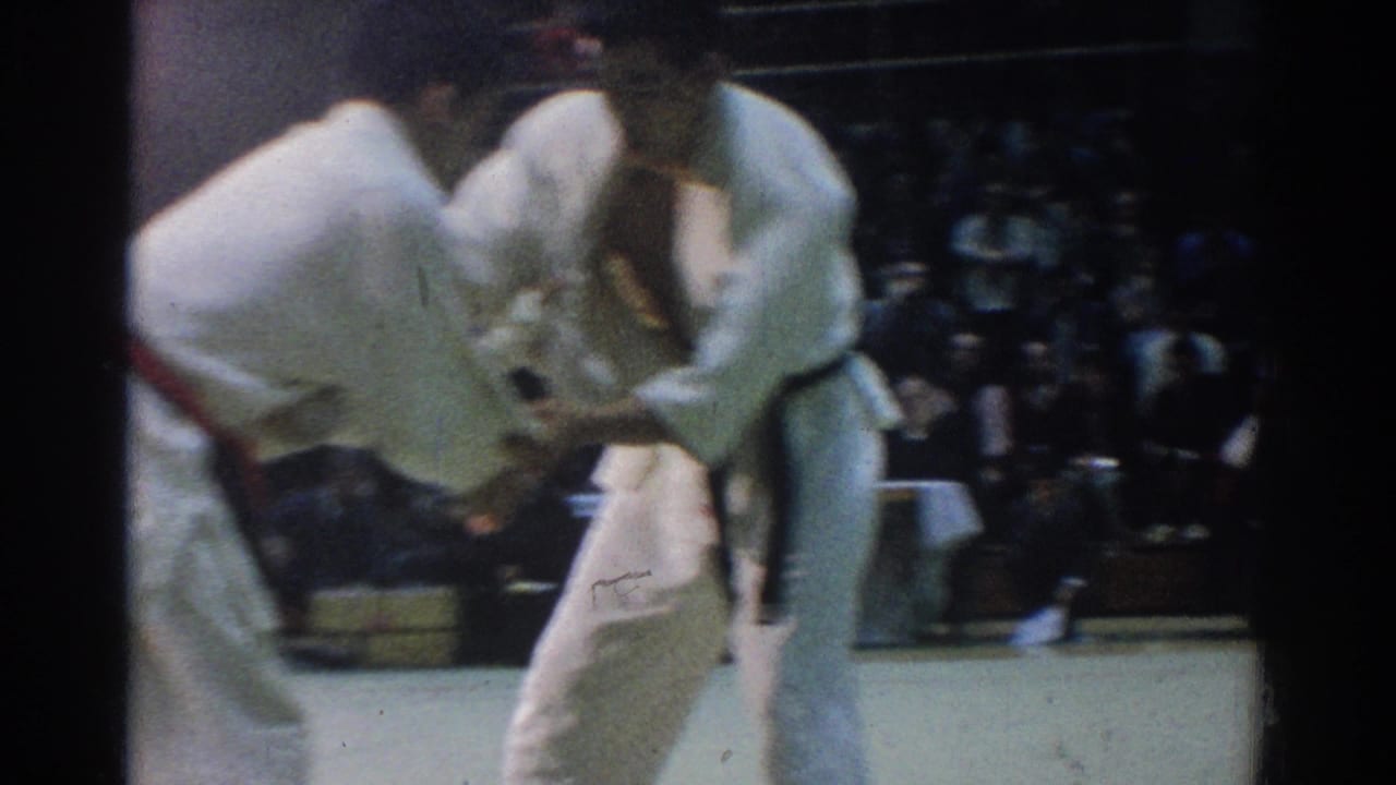 Two judokas in white gis grapple on an indoor mat before a seated audience.