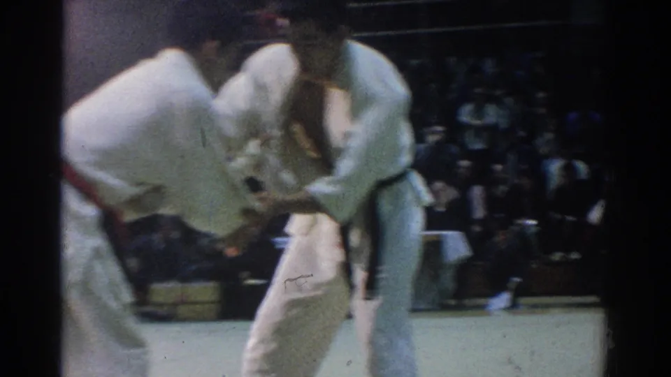 In 1966 West Covina footage, two male judokas in white gis lean into a grip exchange on a mat with spectators behind them.
