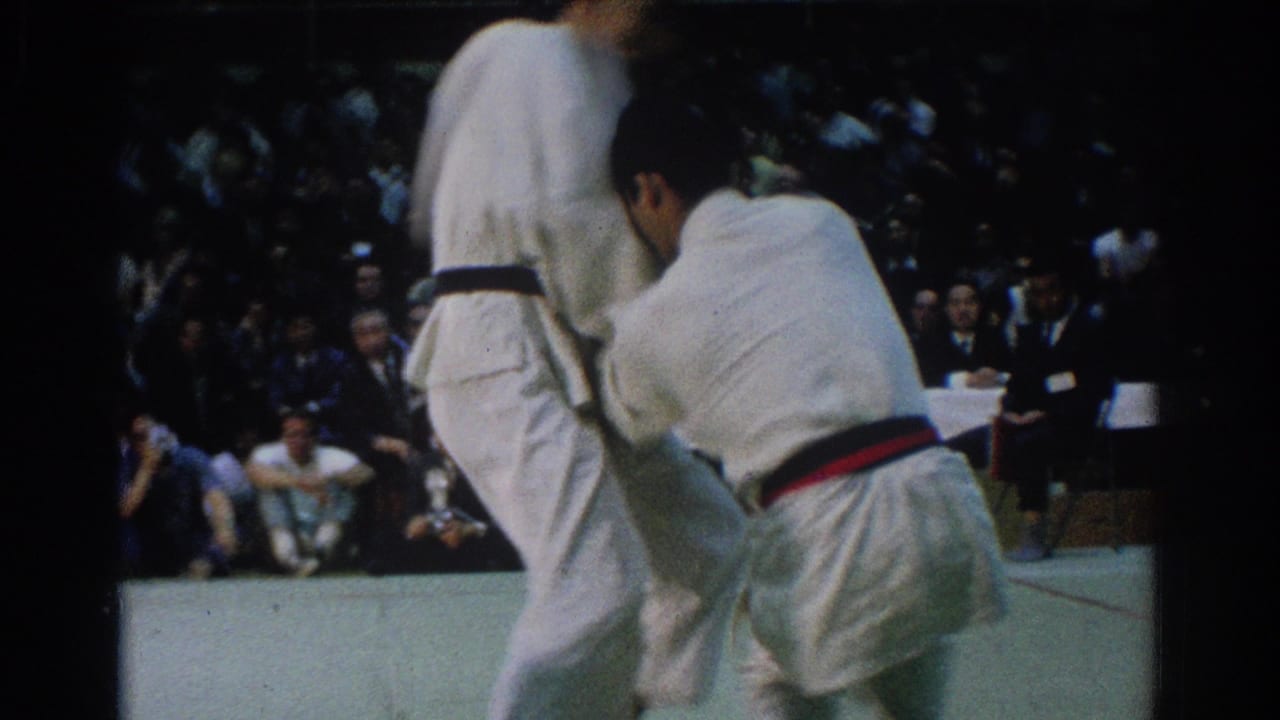 The red-belt competitor holds position beside a black-belt opponent during the 1966 West Covina judo match, with audience members seated in back.