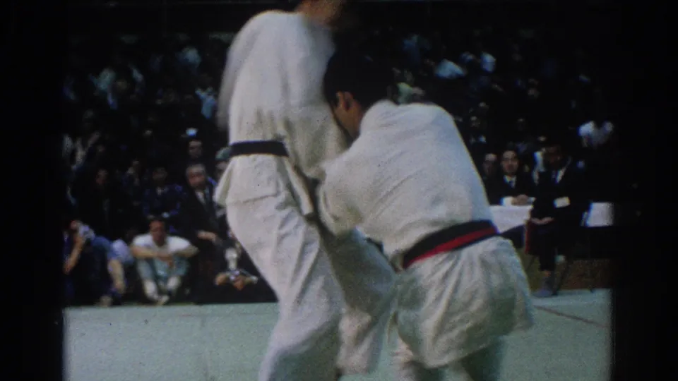 The red-belt competitor holds position beside a black-belt opponent during the 1966 West Covina judo match, with audience members seated in back.