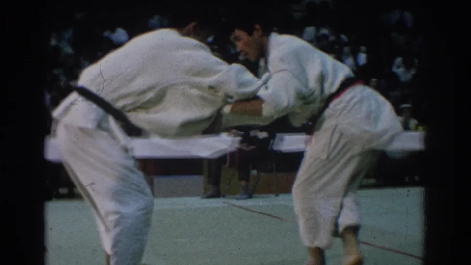 Two judokas in white gis maintain sleeve and lapel contact on the indoor mat, framed from a close home-movie spectator angle.