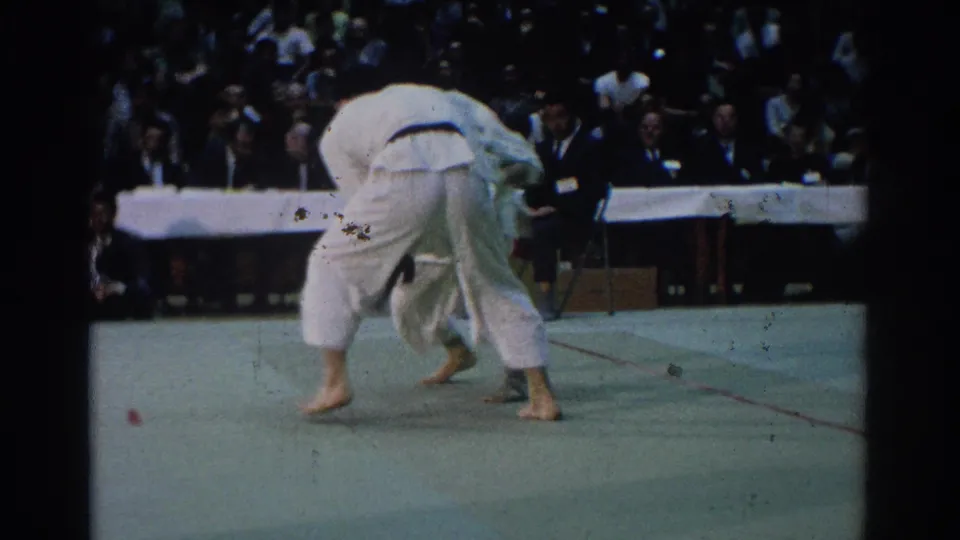 A competitor in a white gi bends over another judoka during a throw or hold attempt, with judges and spectators visible in the background.