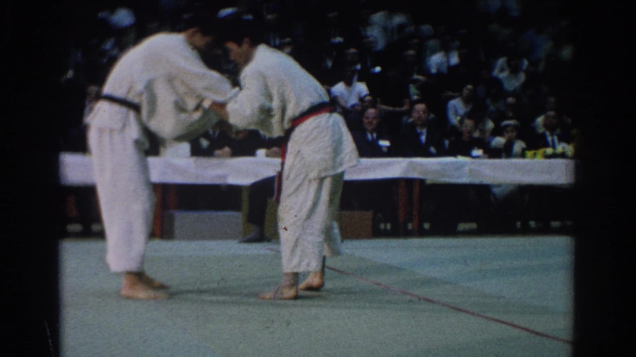Two martial arts competitors in white gi grapple on a green mat before a seated audience.