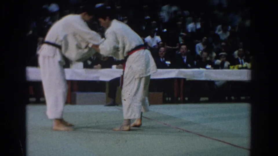 At a 1966 West Covina tournament, two men in white gi and colored belts begin grappling on a green mat as spectators watch.