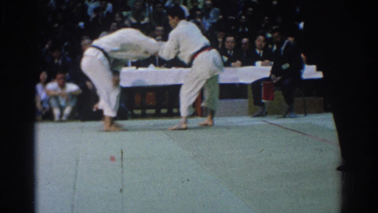 Two competitors lean into a grapple while judges sit behind a white-covered table.