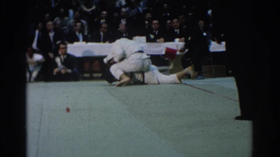 One martial arts competitor appears grounded on the green mat while the other stays above him, with officials and spectators watching.