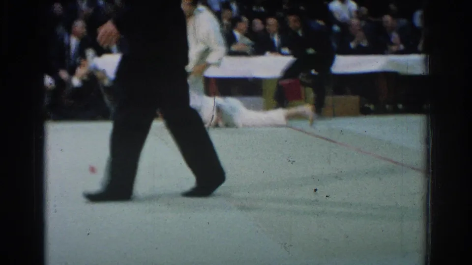 A man in a dark suit crosses the foreground, partially blocking the view as a competitor in a white gi gets up on the mat.