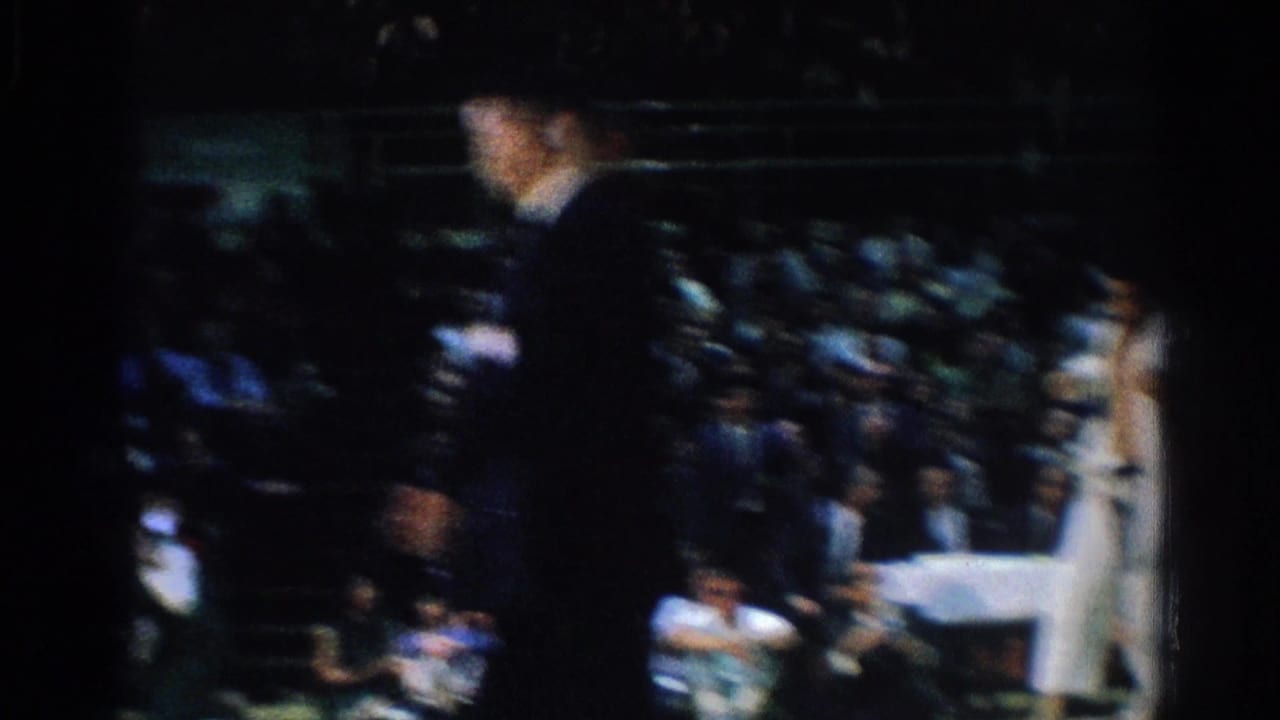 A suited official and judo competitors are seen near a mat before a crowd.