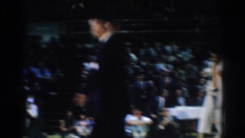 In West Covina in 1966, a suited official and white-uniformed judo competitors appear beside a crowded indoor match area.