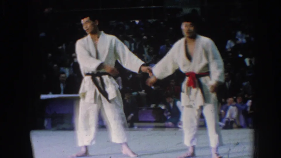 Two judo competitors, one wearing a black belt and one wearing a red belt, shake hands on a West Covina mat in 1966.