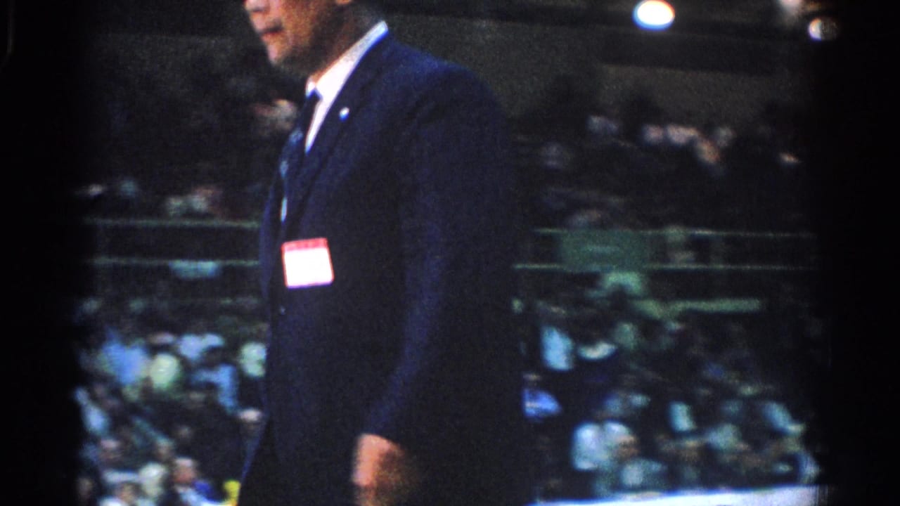 Suited referee with name tag walking past spectators in bleachers.