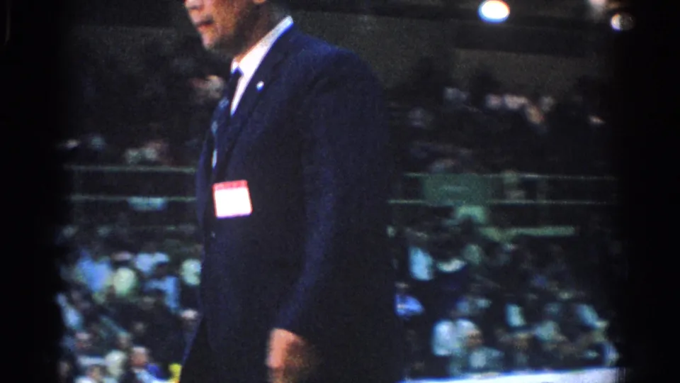 In 1966 West Covina footage, a suited referee with a name tag moves across the foreground as spectators sit in gym bleachers behind him.