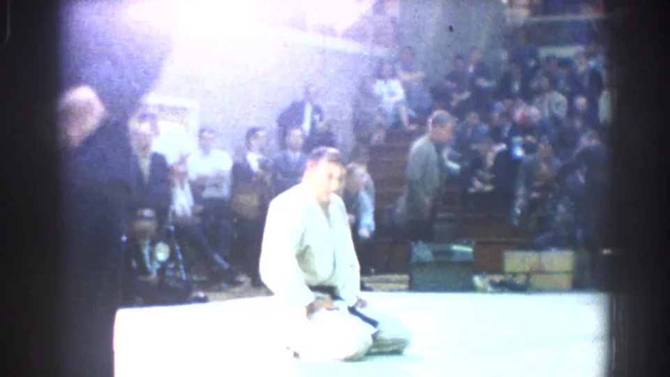 A karate competitor in a white gi kneels on the bright mat as officials and spectators watch from the gymnasium background.