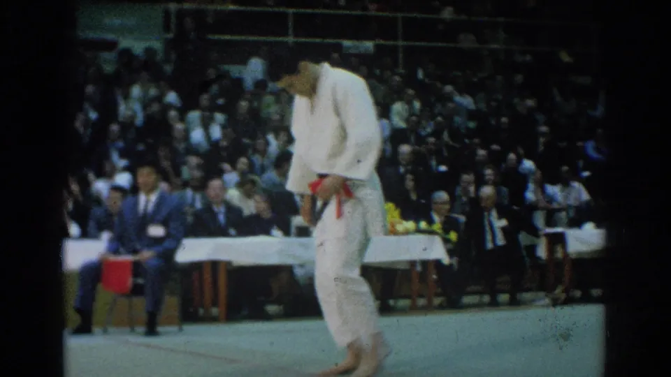 A karate fighter in a white gi stands on the gym mat, adjusting or holding a belt while the audience watches from behind.