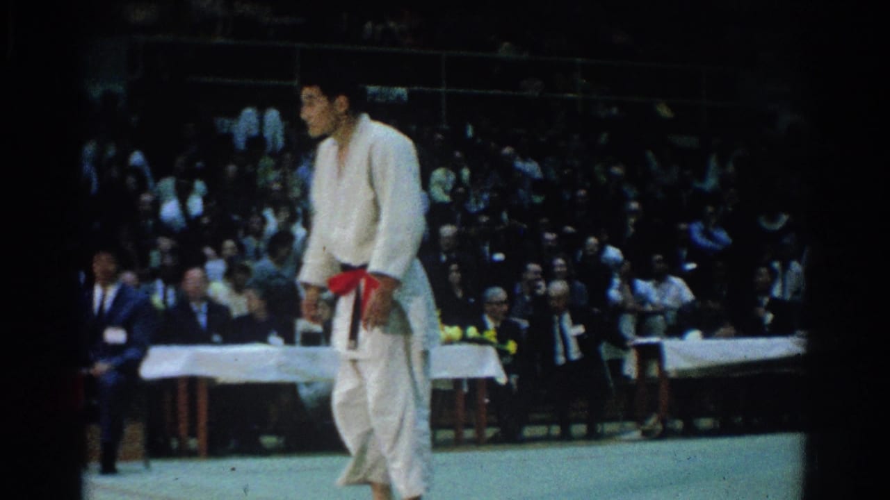 Judo competitor in a white gi with a red belt stands on a mat before seated spectators.