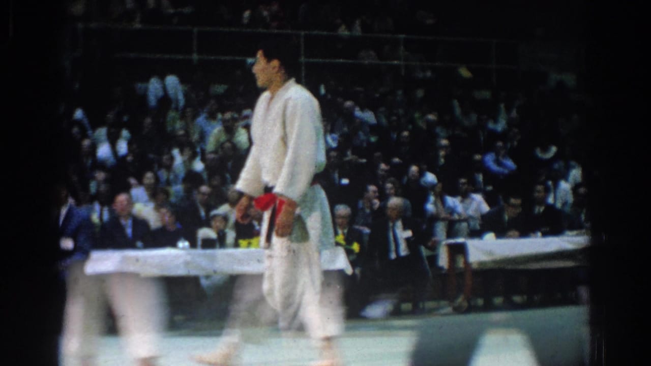 1966 West Covina indoor judo footage with a red-belt competitor crossing the mat and spectators seated beyond the judges table.
