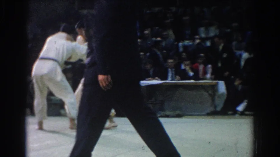 Two judo competitors in white gi uniforms grapple during the West Covina match, with a referee nearby and spectators in the background.