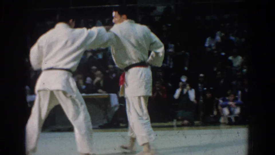 The match continues with two white-gi competitors facing off on the indoor mat before a crowded spectator area in West Covina.