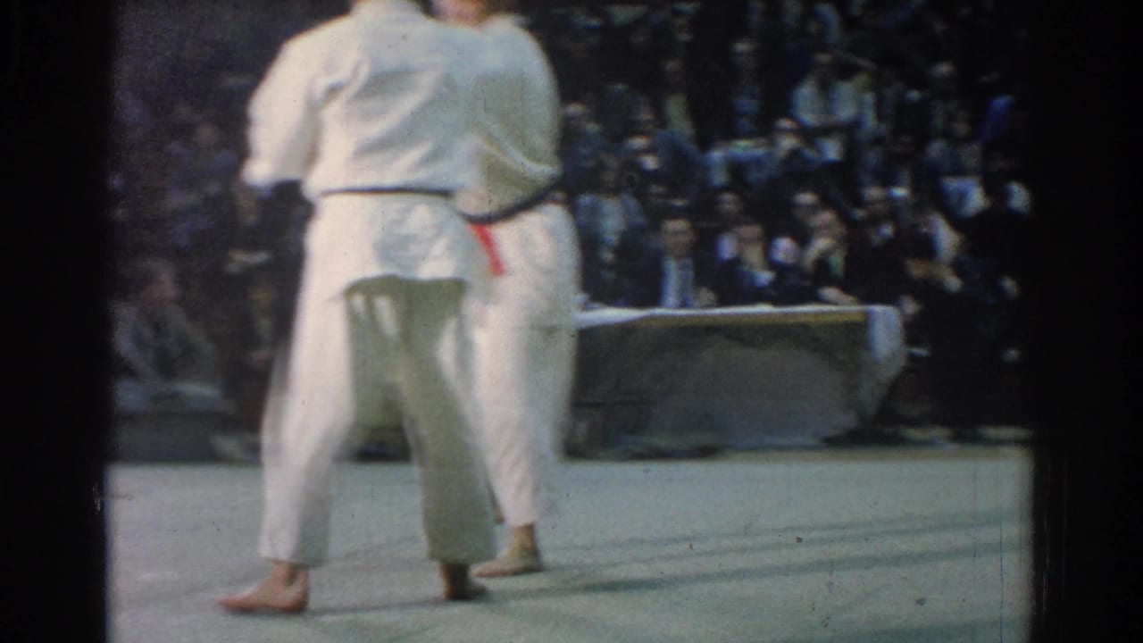 Two martial artists in white gi stand on a mat before spectators.
