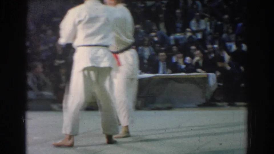 Two martial artists in white gi face each other on a gym mat in West Covina, California, 1966, with spectators seated behind them.