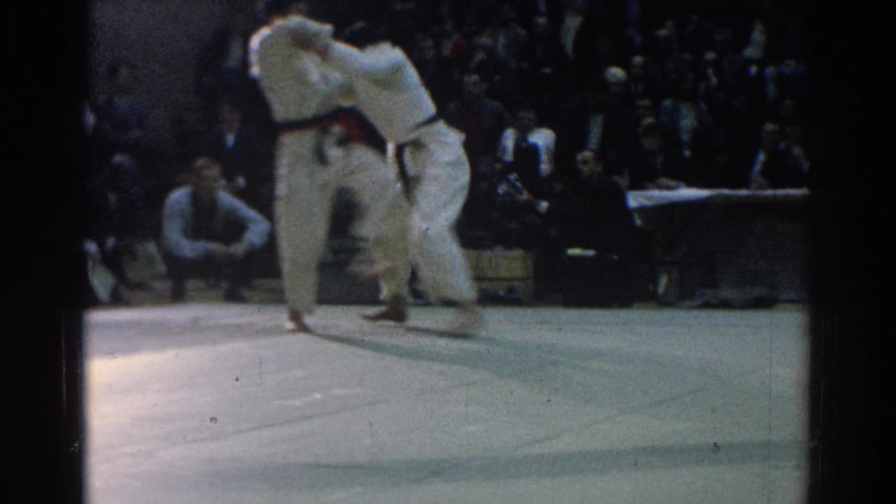 In the 1966 West Covina gym scene, two gi-clad competitors clinch during a judo match as spectators watch from the background.