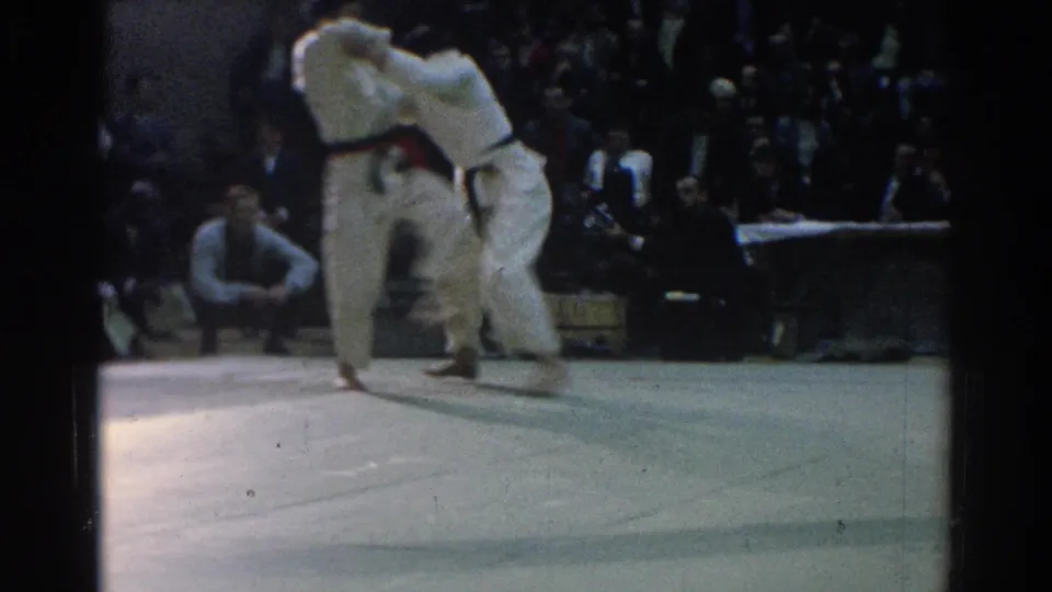 In the 1966 West Covina gym scene, two gi-clad competitors clinch during a judo match as spectators watch from the background.
