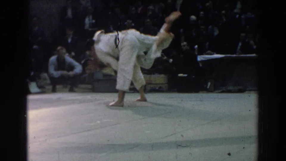 A judo competitor in a white gi lifts and turns an opponent during a throw in the 1966 West Covina gymnasium footage.