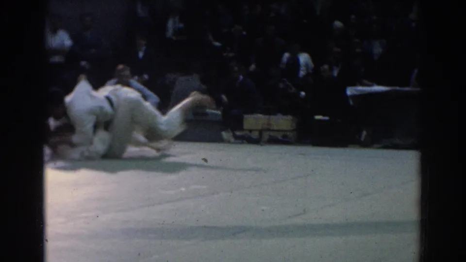 Two martial artists in white gi hit the gym mat after a judo throw, with spectators and a table visible behind them in West Covina, 1966.